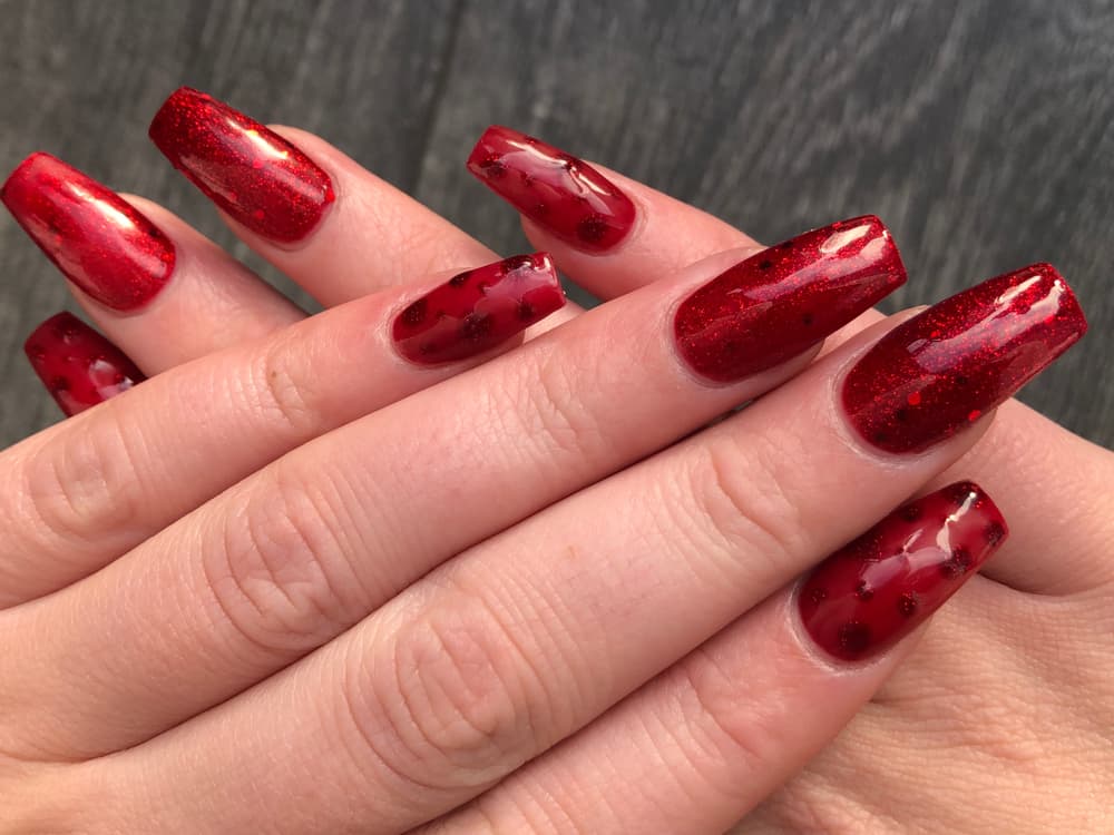 Close-up of hands with a full set of red acrylic nails featuring a glossy finish and glitter accents.