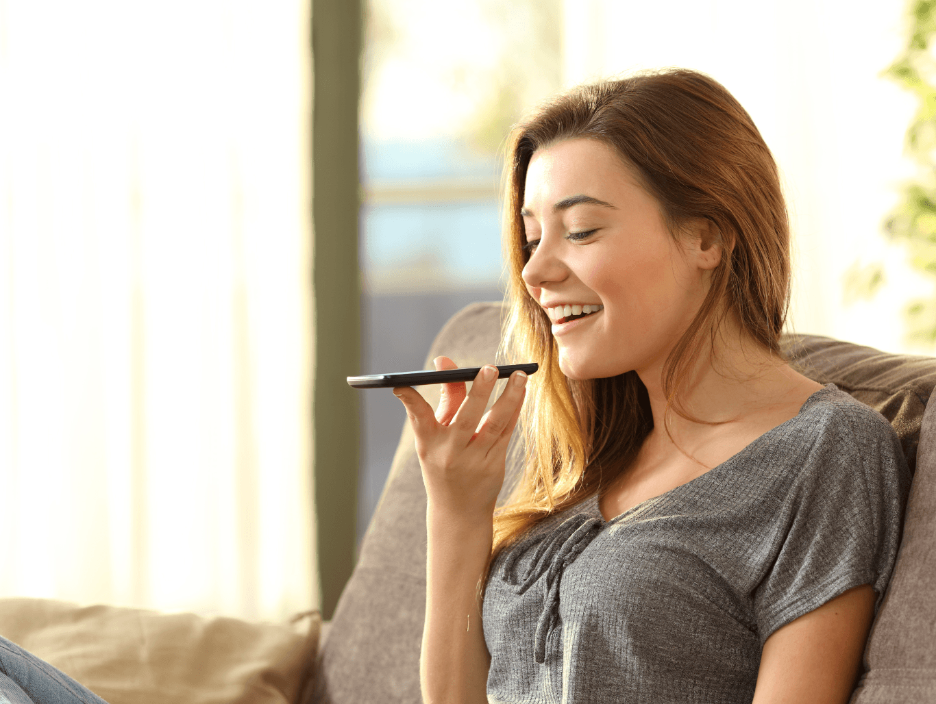 A smiling young woman relaxing on a sofa while recording a voice message on her smartphone in a bright, cosy home setting – symbolising friendly and approachable communication.
