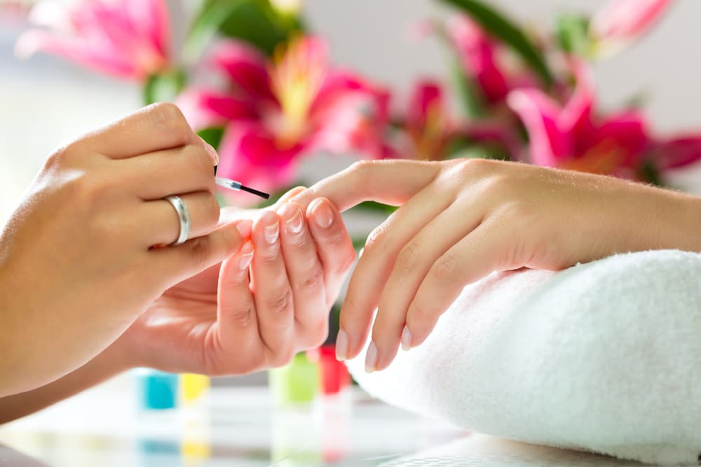Close-up of a beauty therapist applying nail polish during a luxury manicure, with fresh flowers in the background.